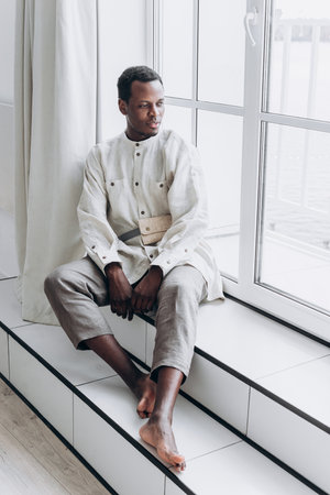 Black Man in Minimalist Linen Clothing Sitting by a Bright Windowの写真素材