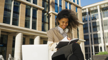Smiling Young Woman Working on Laptop and Taking Notes Outdoorsの写真素材