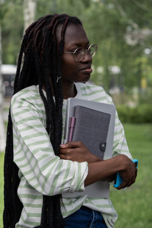 Young African American woman with dreadlocks and glasses holding laptop and books, standing...
