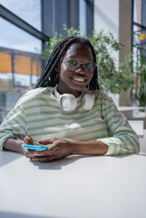 Smiling African American Woman with Braided Hair and Headphones Using Smartphone in Modern cafe