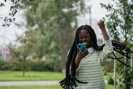 Joyful young African American woman with long braids celebrating success while looking at smartphone in parkの写真素材