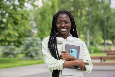 Smiling African American Woman with Braided Hair Holding Laptop and Notebook in Urban Parkの写真素材