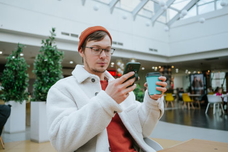 Man in warm coat and beanie using smartphone and holding coffee cup in a modern indoor cafe with...