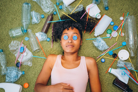 Young woman lying on carpet grass surrounded by plastic bottles and straws, highlighting the issue...