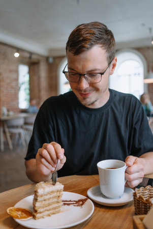 Man enjoying coffee and layered cake at a cozy cafe. Male breaks off piece of cake with dessert spoon.の写真素材