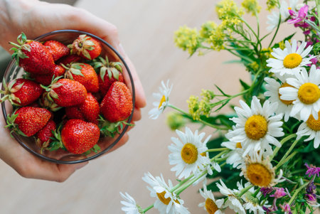 Woman hands holding a bowl of fresh strawberries next to a bouquet of daisiesの写真素材