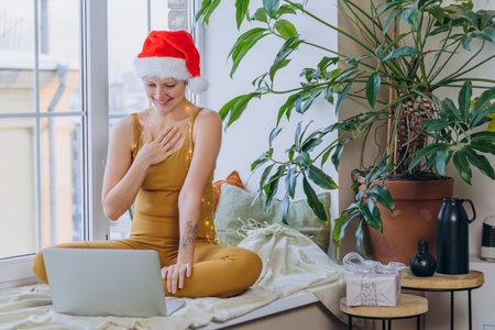 Woman in Santa hat practicing yoga and meditation by window with laptop during Christmas seasonの写真素材