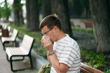 Man sneezing into tissue while sitting on a bench in a park, wearing striped shirt and glassesの写真素材