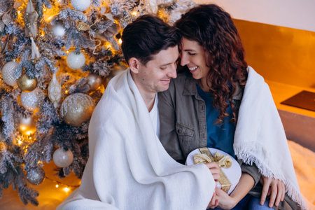 Happy couple sitting by Christmas tree wrapped in blanket, holding gift, cozy holiday sceneの写真素材