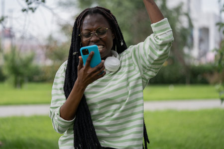 Excited african american woman rejoices and celebrating good news on smartphone outdoors