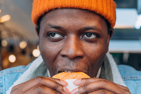 Close-up of African-American man in orange hat and denim jacket biting burgerの写真素材