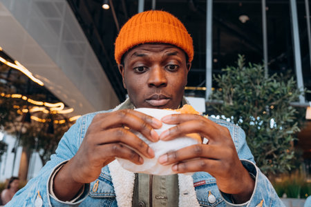 African American man with surprised look holding burger in hands while sitting in food court of shopping centerの写真素材