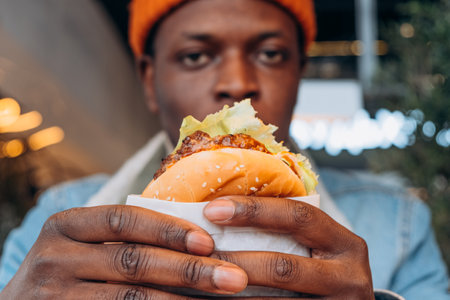 Close-up of black man holding juicy burger with lettuce and sauce, focusing on foodの写真素材