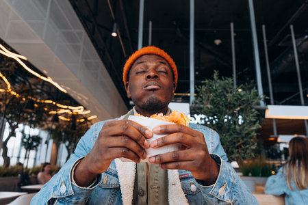 Black man enjoying delicious burger in modern restaurant with eyes closed with pleasureの写真素材