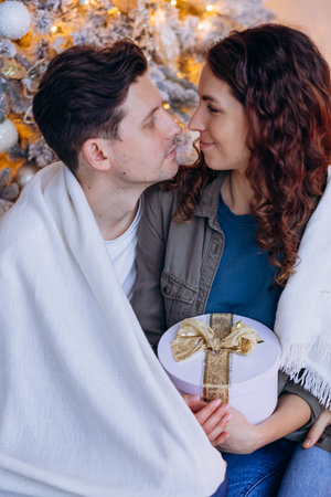 Lovers wrapped in blanket sitting at Christmas tree and looking into each others eyes, holding wrapped gift in handsの写真素材