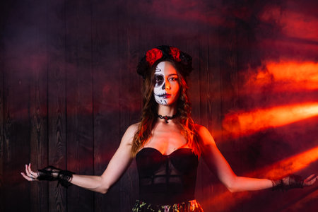 Spooky woman with artistic makeup poses in dark room surrounded by red smoke to celebrate the day of the deadの写真素材