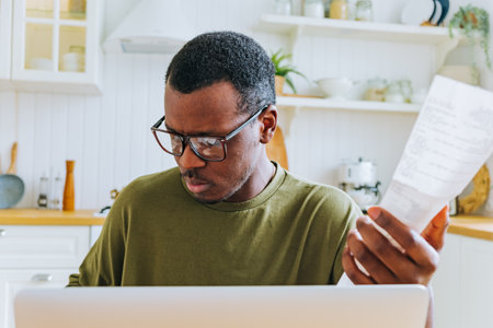 Focused young african american man in glasses working from home, managing monthly expenses and checking bills in the kitchenの写真素材