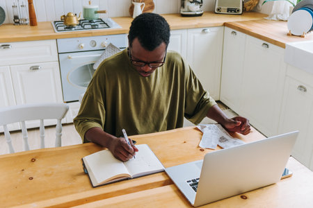 Young african american business owner manages finances and plans budget from home, using a laptop at his kitchen tableの写真素材