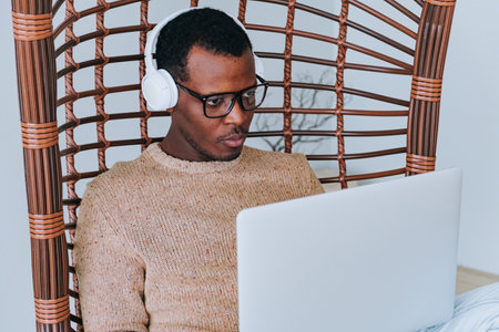Young professional with headphones is focused on work, sitting on a cozy swing chair with his laptopの写真素材