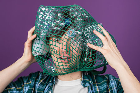 Man struggling to remove a green plastic bag filled with empty bottles from his head while expressing concern about plastic pollutionの写真素材