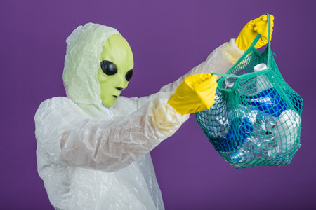 Alien in protective gear holding bag of plastic bottles promotes recycling and environmental consciousnessの写真素材