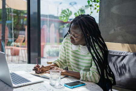 Young professional working remotely in a cafe, taking notes in a notebook and using a laptop while dressed casually with headphones around her neckの写真素材