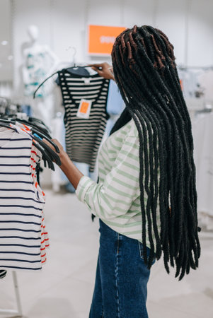 Young african american woman with dreadlocks shopping in a trendy fashion boutique, choosing a stylish black and white striped shirt on a hanger. Focus on price tag for sale optionsの写真素材