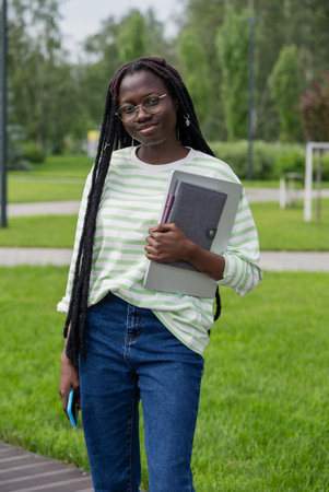 Confident young african american woman walking through campus park with laptop, notebook, and smartphone for online class, surrounded by lush greenery, embracing technology in educationの写真素材