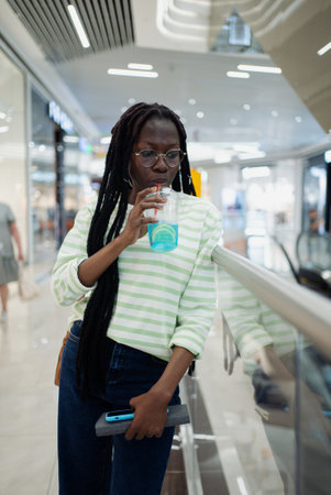Young woman pauses her shopping trip to enjoy a refreshing drink indoors while checking her phone, with the modern interior reflecting in the window behind herの写真素材