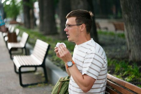 Young man in a city park battles allergies and sneezing caused by pollen, highlighting the need for...