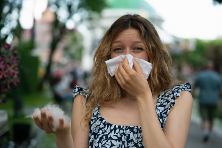 Determined young woman with allergies blowing her nose in a park filled with poplar fluffの写真素材