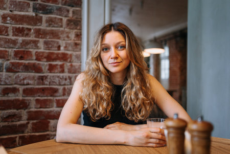 Young woman with long blonde hair sits at a restaurant table, holding a glass of water and waiting, wearing a black topの写真素材