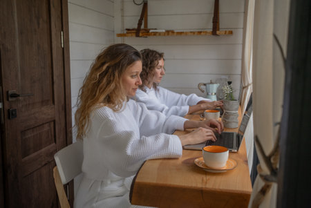 Two women, clad in comfortable white robes, work intently on their laptops in a well-lit wooden cabin, with steaming cups of coffee at their side.の写真素材