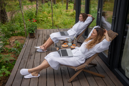 Two women in white bathrobes are relaxing on wooden deck chairs, with laptops and coffee on a deck surrounded by a lush forestの写真素材