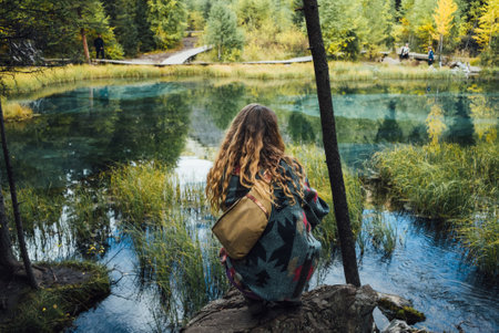 A woman with long, curly hair gazes out at a lush forest pond, reflecting the beauty of the tranquil surroundings, with a rustic bridge and fellow explorers in the background.の写真素材