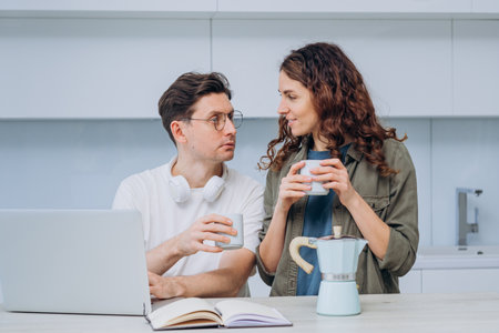Young couple taking a break from work to savor a cup of coffee together in their cozy kitchen. Both are smiling and gazing at each otherの写真素材