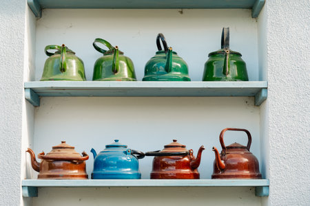 Vintage teapots in various colors and finishes are neatly arranged on a blue shelf. The collection evokes a sense of nostalgia and warmth.の写真素材