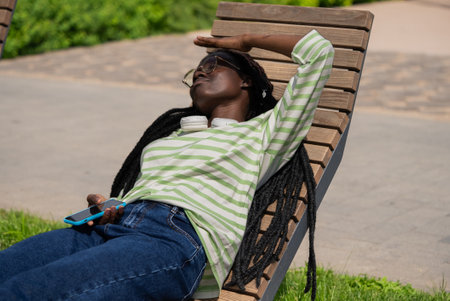 Young woman relaxing on bench in park shielding eyes from sunの写真素材