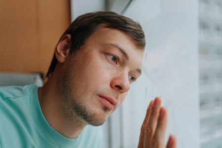 Young man suffering from depression looking out the windowの写真素材