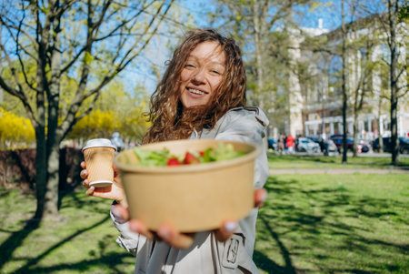 Young woman happily eating a healthy meal in a lush park, holding a salad bowl and coffee cup, smiling in the sunny outdoors. Her curly hair blows in the breeze, creating a positive urban vibeの写真素材