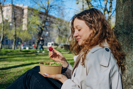 Young woman with curly hair savors a healthy lunch in a city park, enjoying each bite in the sunshineの写真素材