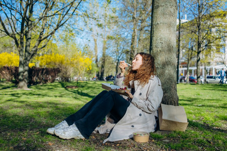 Young woman enjoying a healthy lunch under a lush tree in a serene park on a sunny spring day, savoring her meal in natureの写真素材