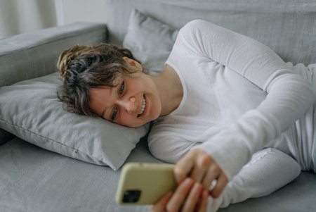 Smiling woman using smartphone lying on sofa at homeの写真素材