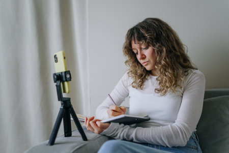 Influencer creating content at home writing on notebook filming with smartphoneの写真素材