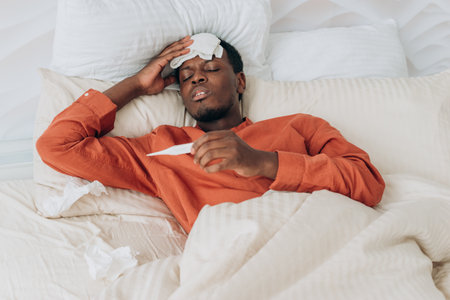 A man lies in bed with a thermometer, a cool cloth on his forehead suggesting illness, in a scene of home health care.の写真素材