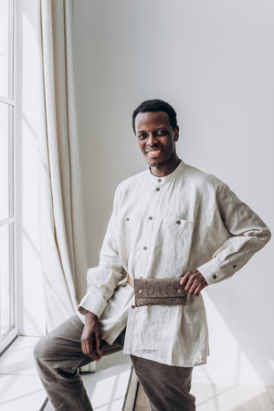 Smiling black man wearing traditional linen clothes posing by windowの写真素材