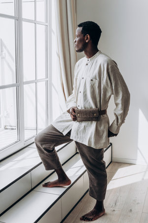 Stylish black man wearing linen clothes and fanny pack looking out windowの写真素材