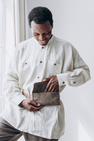Smiling black man opening eco-friendly felt bag by windowの写真素材