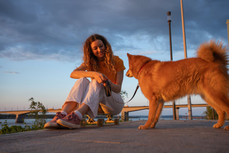 Young woman sitting on skateboard and playing with dog by river at sunsetの写真素材