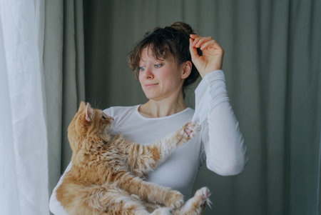 Woman holding ginger cat while adjusting hair near windowの写真素材